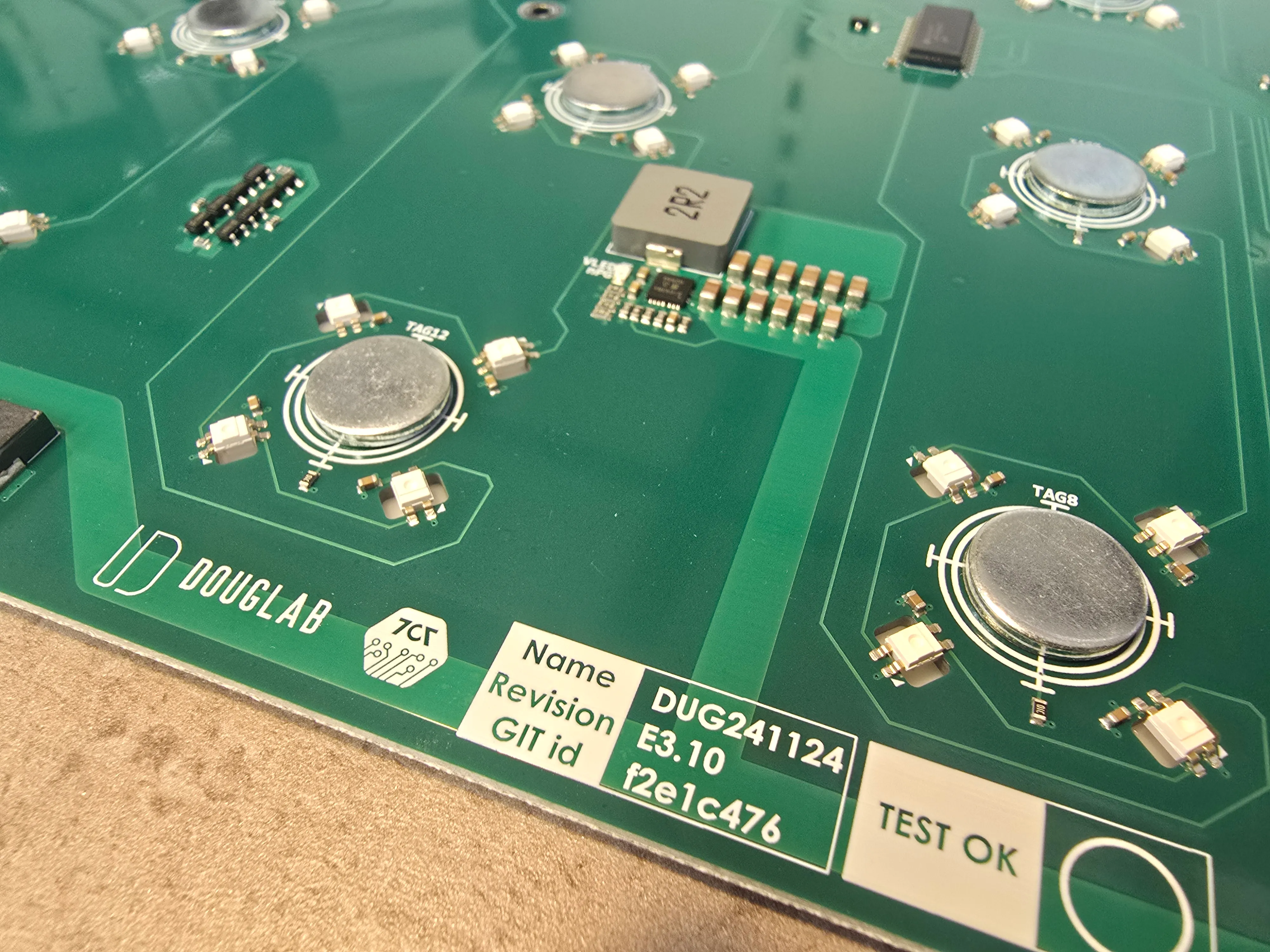 Close-up of a green MagMAGI main board with round badge positions surrounded by LEDs, the DougLab and 7c7 logos and a TEST OK label