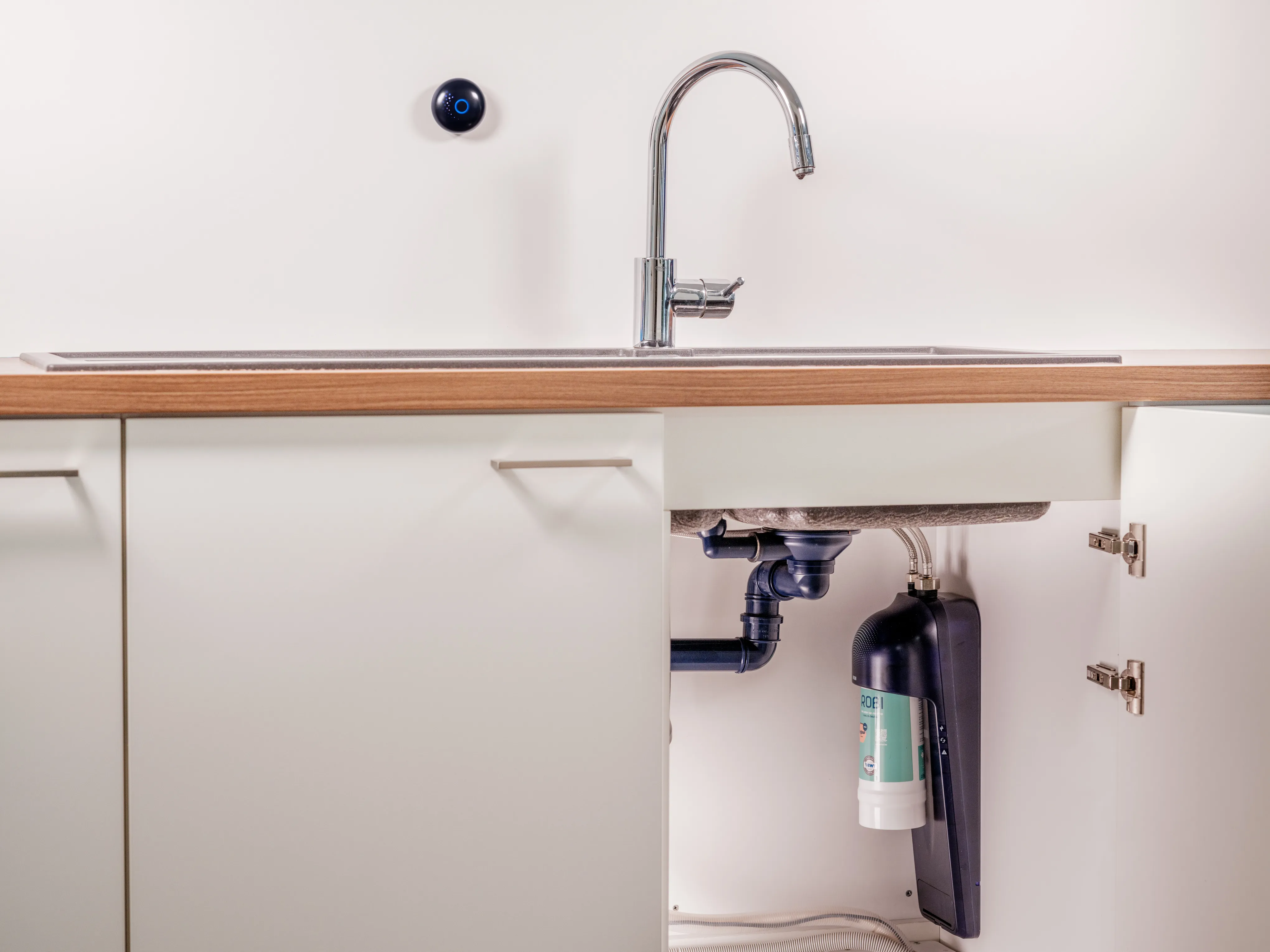 ROBI filter unit mounted under a kitchen sink, with its wireless button on the wall beside the tap
